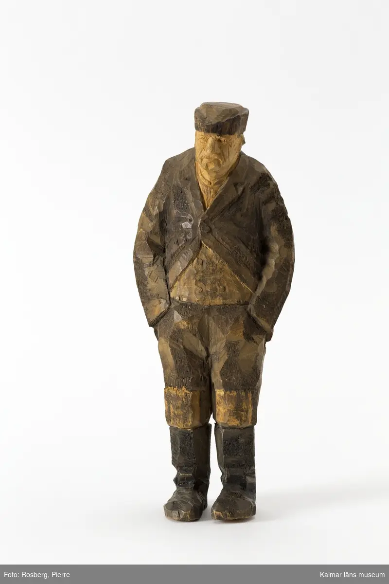 KLM 45932:2 Skulptur, av trä, målad. Figur i form av en stående man. Han har båda händerna i byxfickorna. Klädd i huvudbonad, jacka, väst och byxor med lappar på knäna, samt stövlar. Signerad på ryggen, H.C. Målad i bruna nyanser och svart. Tillverkad av Helge Rugland, f. Carlsson.