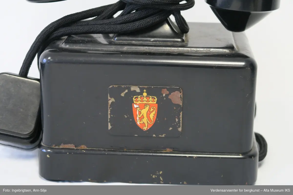 Prismeformet boks med bæreklo på toppen og telefonrør. Emblem med det norske riksvåpenet på framsiden av boksen. På siden er det festet en strømledning med støpsel i enden.