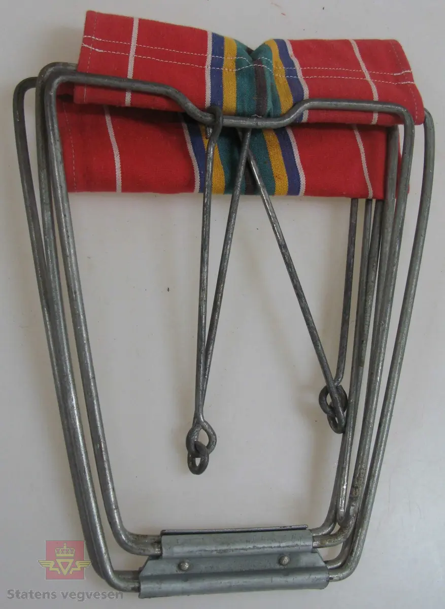 Enkel campingstol med rammeverk av stål og trekk av tekstil. Trekket er flerfarget med striper, hovedfarge er rød. Stolen kan pakkes helt ihop når den ikke er i bruk.