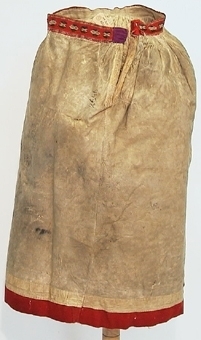 Kjol av fårskinn med hårsidan inåt och röd klädeslist nertill. I midjan vävt band i vitt och grönt mönster. Kjolen är sydd av fyra våder samt har stripade rynkor vid linningen.