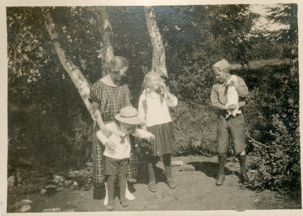 Tre barn og en kvinne står utendørs i en hage eller park. Den ene gutten har på seg en  sepeiderdrakt og holder en hvit kanin.