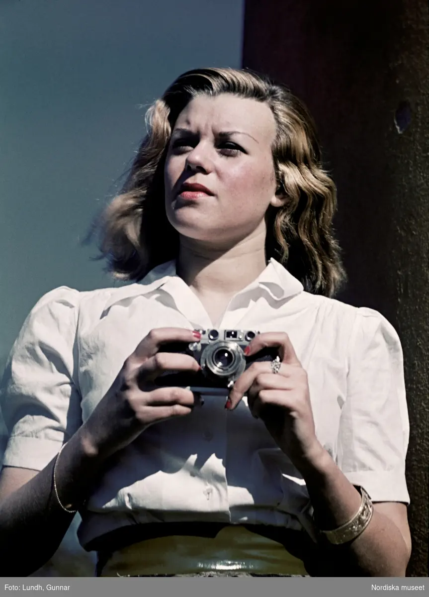 Kvinna i vit blus som bär smycken håller i en småbildskamera.