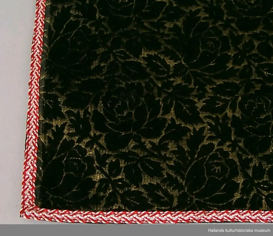 Rektangulär duk av mönstrat sammetstyg. Mönstret är bestående av svarta rosartade blommor på guldfärgad botten. Baksidan svart, med visst genombrott av samma mönster. Duken kantad av rödvitt, smalt band.