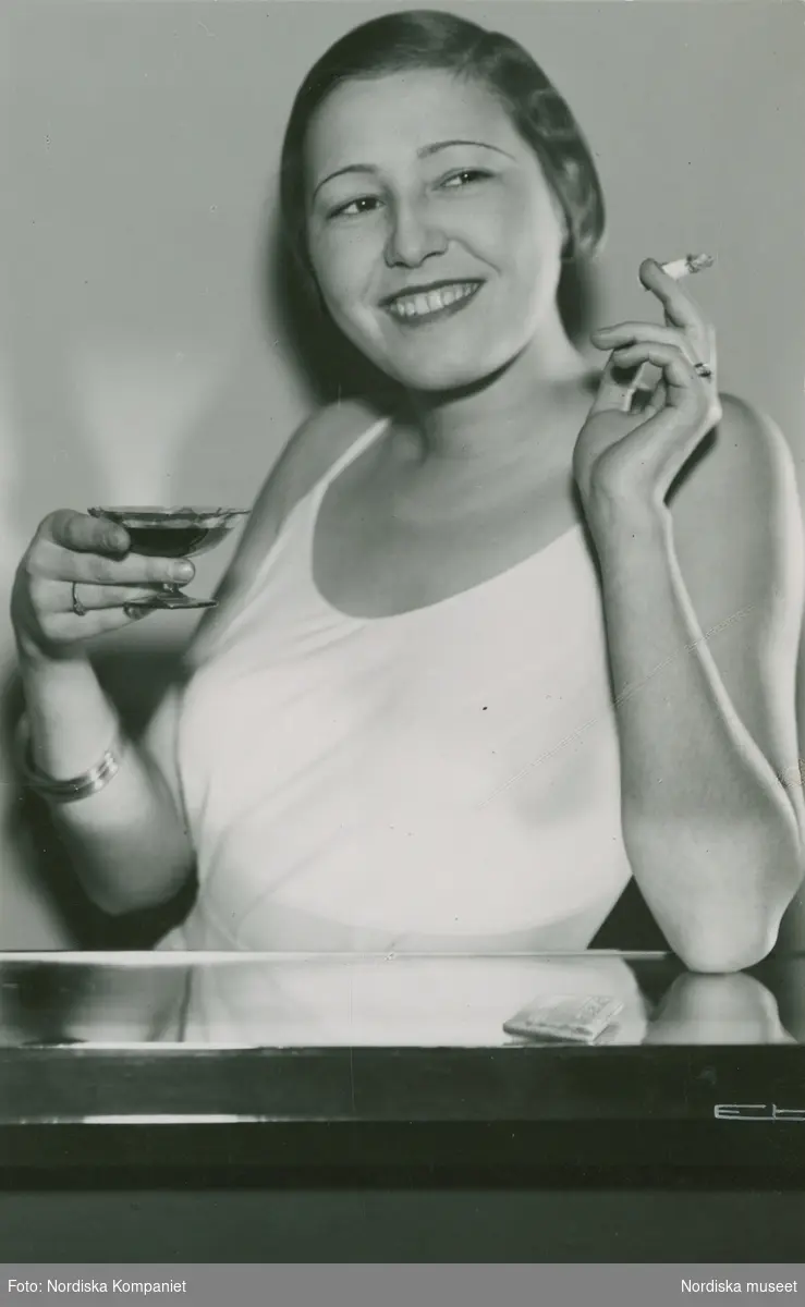 Leende kvinna i slät, vit klänning med tunna axelband. Cigarett och coctkailglas. Nordiska Kompaniet.