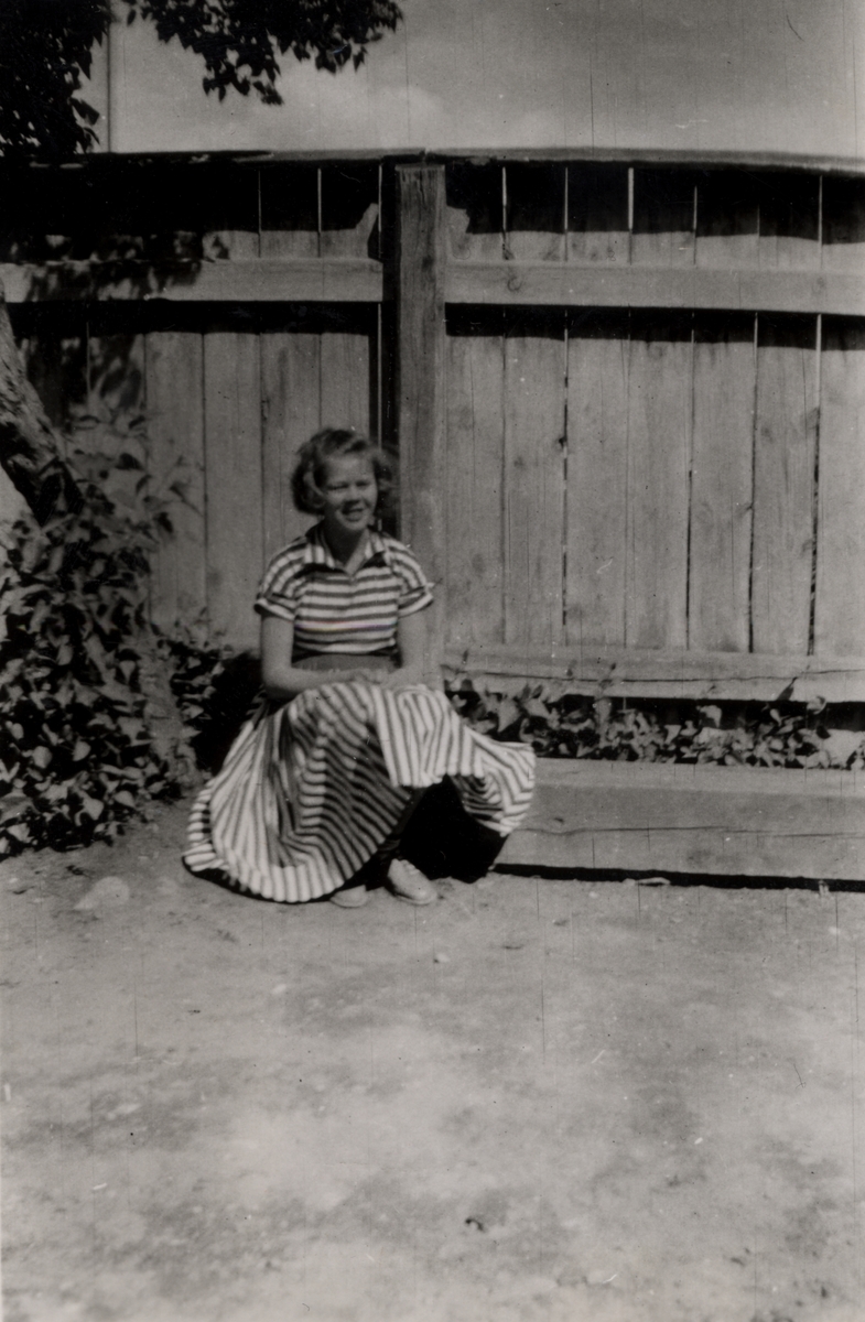 Bilden visar Inger Lilja som 16- åring, sittandes i randig klänning med ett plank i bakgrunden. Kortet är taget vid Drottninggatan 15.