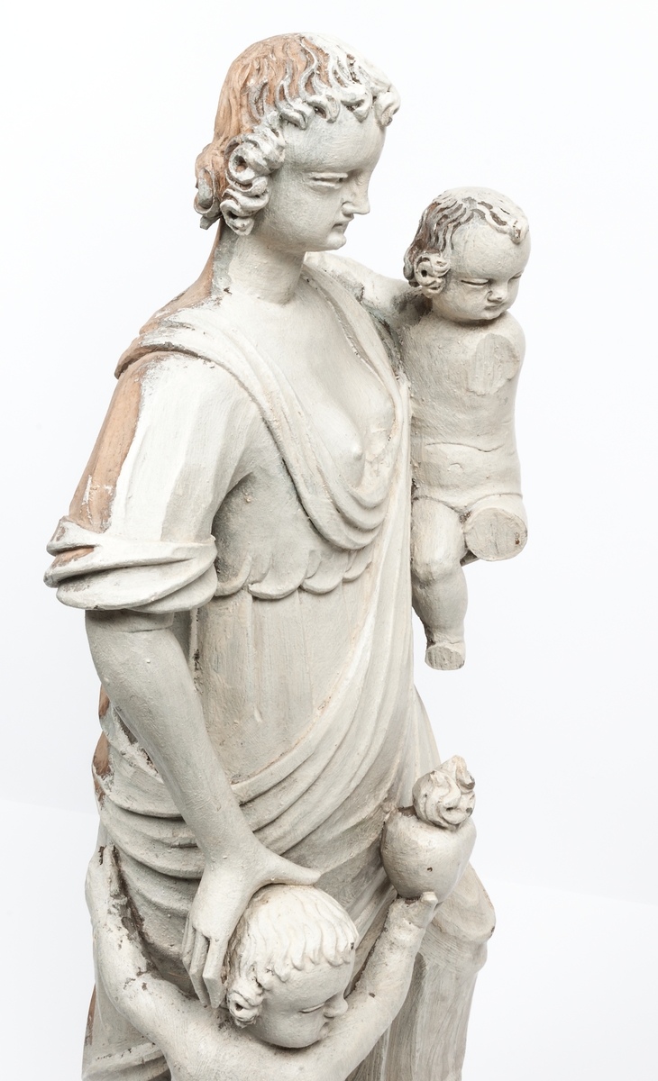 Kyrklig skulptur, trä, staty från gamla korskranket i Heliga Trefaldighets kyrka i Gävle, av Ewert Friis omkring 1665. 

Stående kvinnofigur med naket barn på vänster arm samt stående naket barn vid höger sida. Sistnämnda barn har ett brinnande hjärta i höger hand och kvinnans vänstra hand på sitt huvud. Vänster ben är främst, foten avslagen. Sittande barnets ena ben och andra fot avslagna. 
Fyrsidigt, profilerat postament. 
Gråvit färg.
