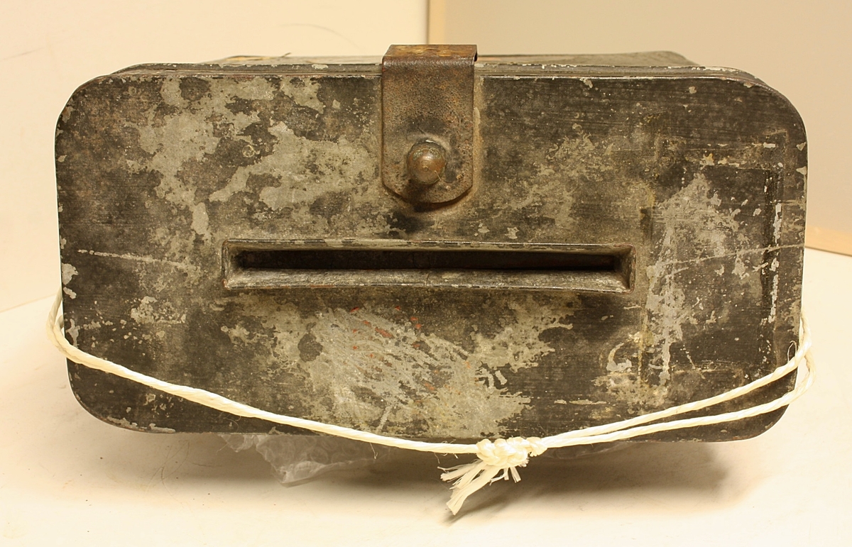 Avrundet, rektangulær boks. Den norske løve på fronten. Rester etter rødt segl. Hvit snor i plast.