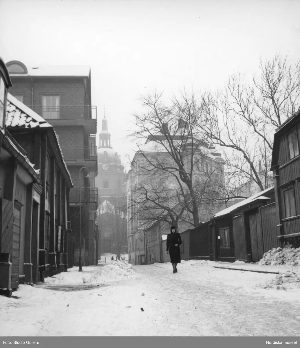 Gatuvy, Mäster Mikaels gata, Södermalm, Stockholm. En kvinna promenerar på den snötäckta gatan, Katarina kyrka i bakgrunden.