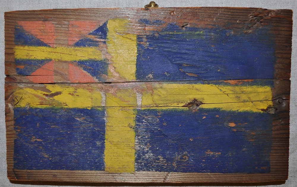 Rektangulär träbit med målad unionsflagga, från ballonghuset på Danskön. Ögla upptill för upphängning. På baksidan text och fastklistrat tidningsklipp om ballonghuset