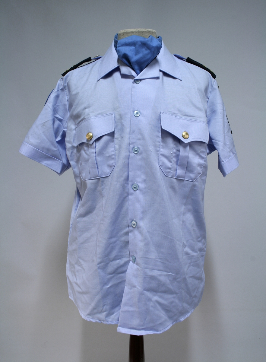 Blå kortermet skjorte med cravat/slips. Skulderdistinksjon for politiførstebetjent.
