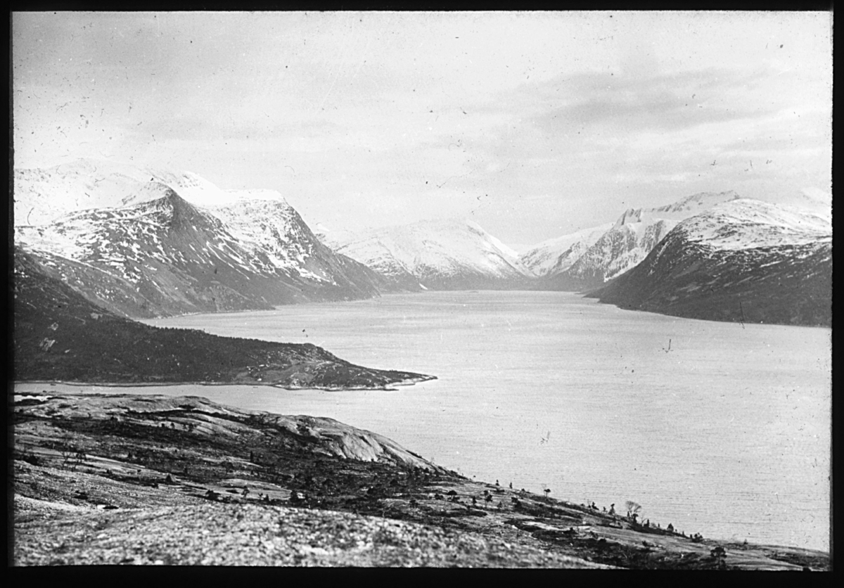 "N.567b). Fra Tysfjord." Landskapsbilde fra Tysfjord. Bildet viser fjorden omkranset av fjell med snøflekker på. I forgrunnen er det bar mark og sparsomt med vegetasjon.