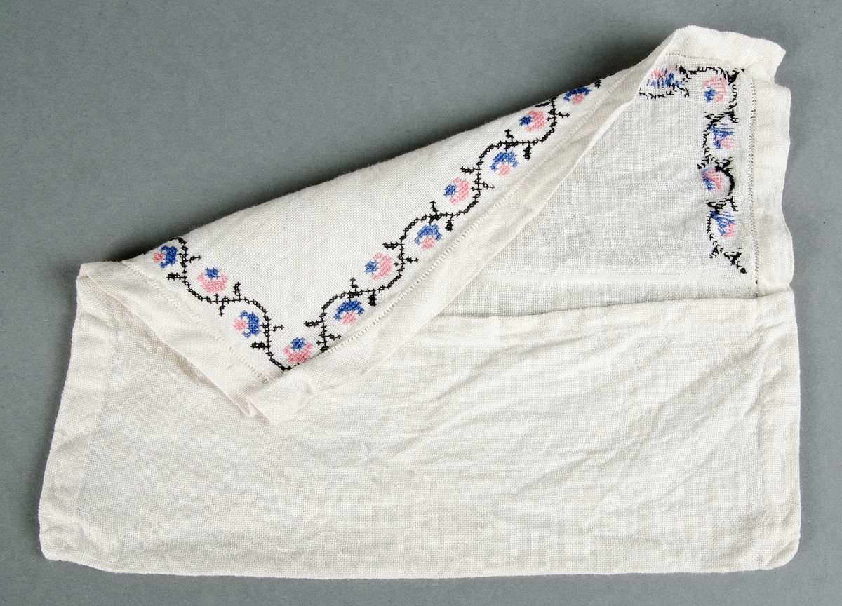 Servettväska av vit linnelärft, handsydd med hålsöm på locket och blombård i korsstygn i svart, rosa och blått. 
Ostruken och skrynklig.

