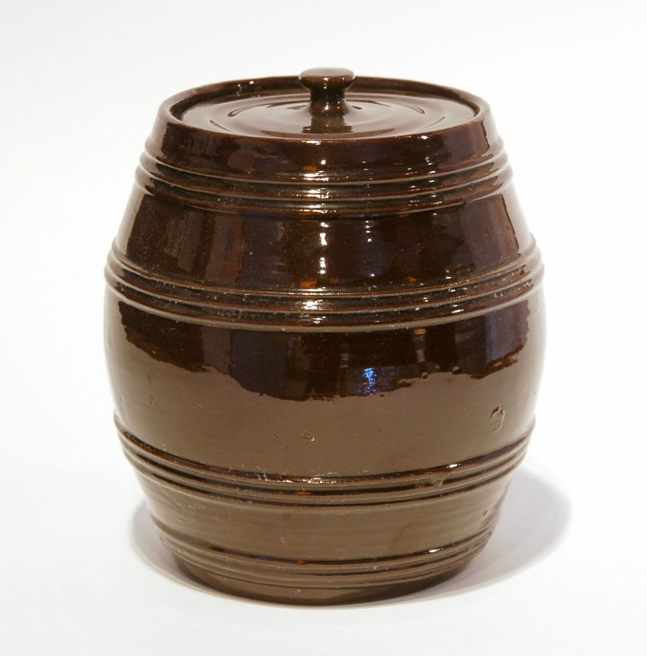 Burk med lock, Bo Fajans, tunnformig, mörkbrun glasyr. Höjd 17,5 cm. 
Prov på fabrikens äldsta tillverkning, 1880-talet.