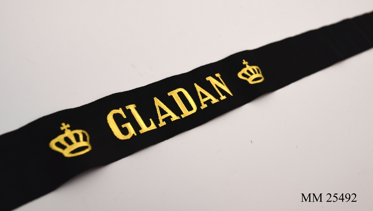 Mössband av svart sidenrips. Guldfärgad text, "Gladan", med två kronor på vardera sida.