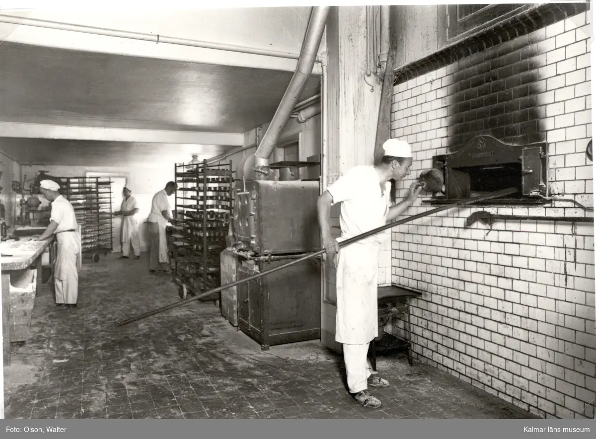 Kalmar Bageriförening. Män som arbetar med bröd, en man står och håller i en brödspade riktad in i en ugn.