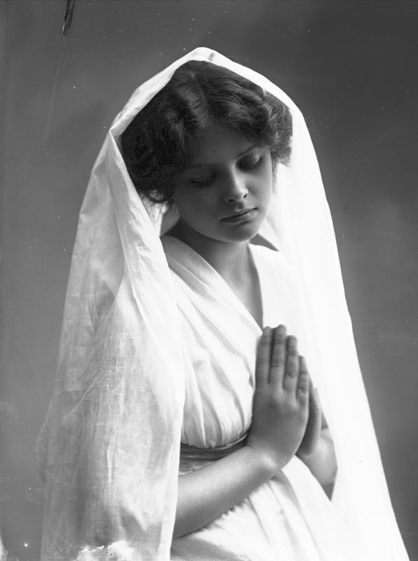 Studioportrett av kvinne i kostyme - hvit kjortel og hvitt slør, som holder hendene foldet. 1908. (Foto/Photo)