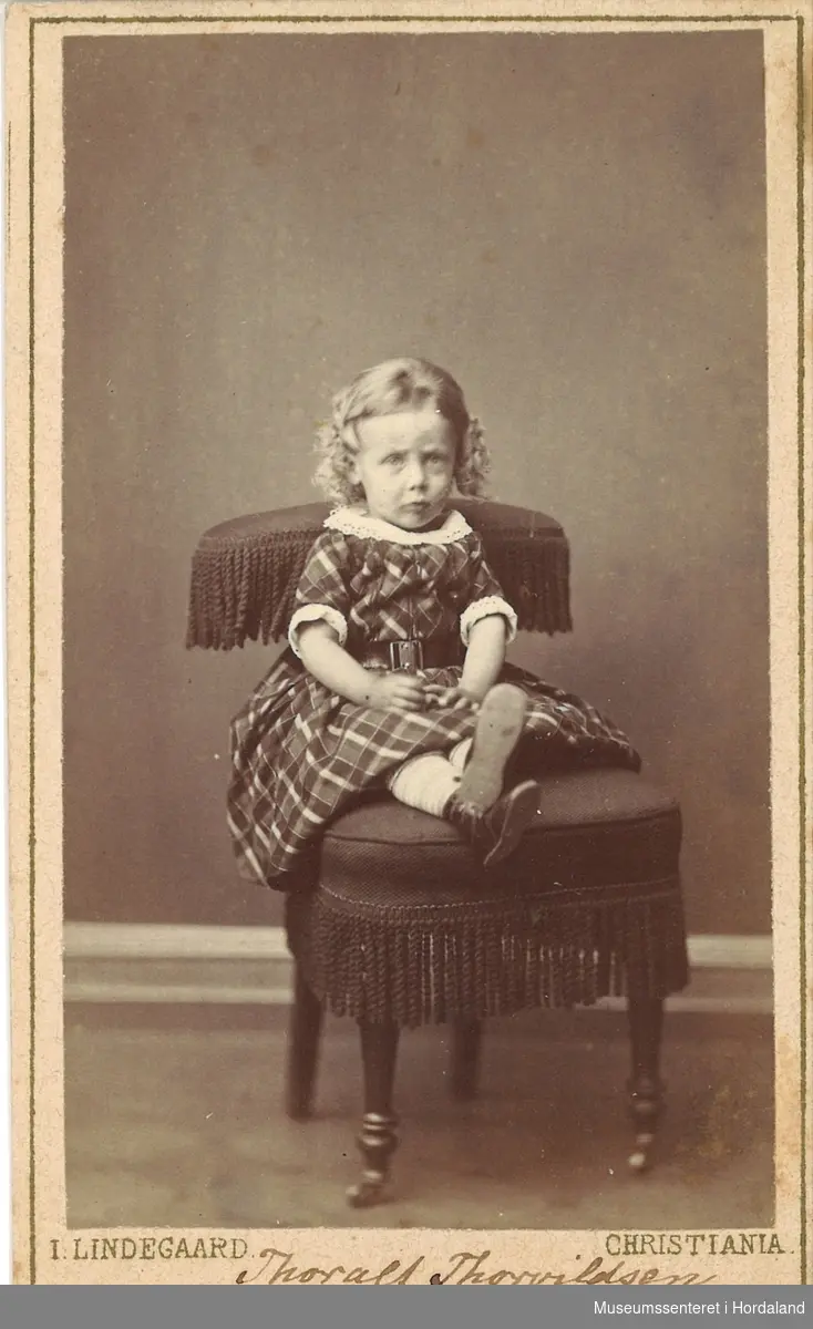 atelierfotografi av ein liten gut med ljose krøllar kledd i ruta kjole som sit på ein stol