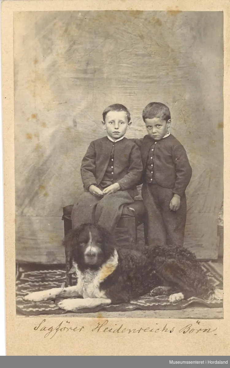 heilfigur fotografi av to  små gutar kledd i jakker, bukser og vest som sitter/står med ein stor St. Berhardshund framfor seg på golvet