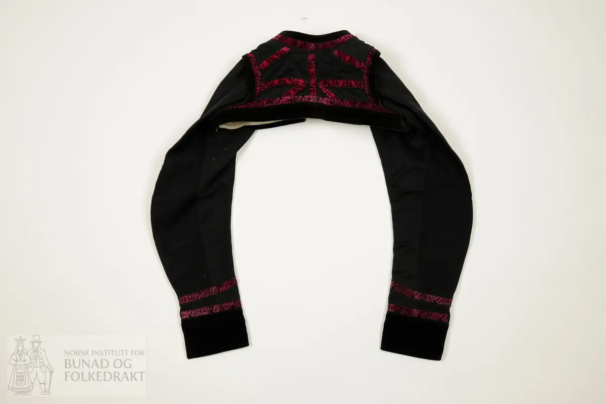 Opentrøye sauma av svart blank glatt klede, lerretsvove. Heilklipt rygg, bandprydd. Oppståande låg krage. Tosaumserme med bryningar. Open ermsliss med 3 stk knappar og knapphol til lukking. Bandpryding av blomstermønstra fløyelsband og svart bomullsfløyel til pryding av krage, framkantar, nedkantar og ermeslissar.  For av kvit litt glissen stiva bomullslerret. Maskinsaum og handsaum.