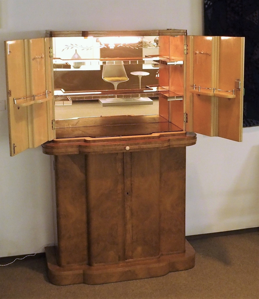 Cockailskap i to høyder med fire dører. Profilert korpus, sokkel, gurtlist og topplist. Mahogny med valnøttfiner; de øvre dørene er finert med lyst satintre på innsiden. Nedre skap har en hylle og en skuff festet øverst. Gurtlisten har en uttrekkbar hylle med ferskenfarget speilglass. Øvre del av korpus har dører som åpner til skap med belysning. Veggen bak, bunnen og to hyller er dekket med ferskenfarget speilglass. Øverst på veggen bak ses geometrisk dekor flankert av bladranker. På hver av dørenes innsider en hylle og gjennomhullet feste for cocktailpinner. Ti originale glasspinner med svaner er bevart.