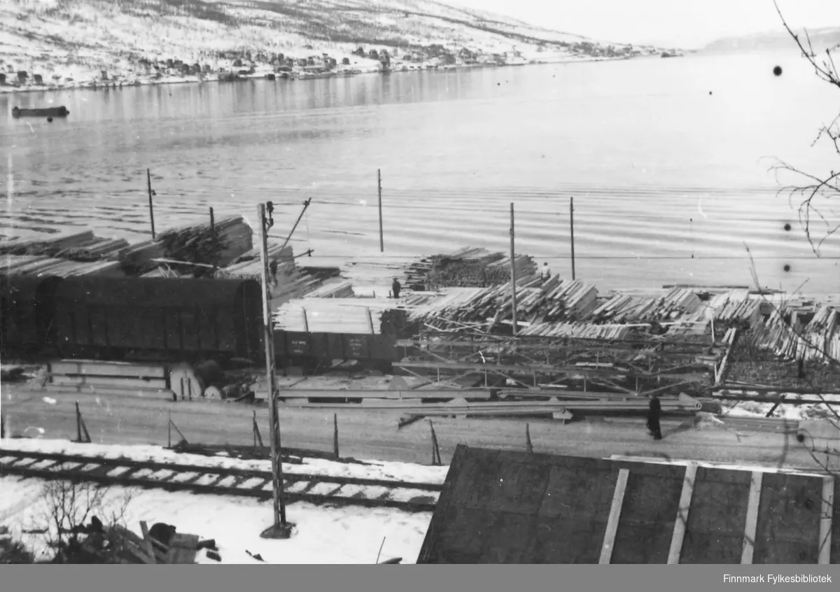 Før og etter 1944, Nordland, Troms. Oversiktsbilde over lagringsplass for trelast. Beliggende på kaia.Stabler med trematerialer ligger stablet opp. Jernbanen passerer iforgrunn. Havet ligger stille og rolig i bakgrunn. På andre siden avsjøen fremtrer noe av bybildet i Narvik.