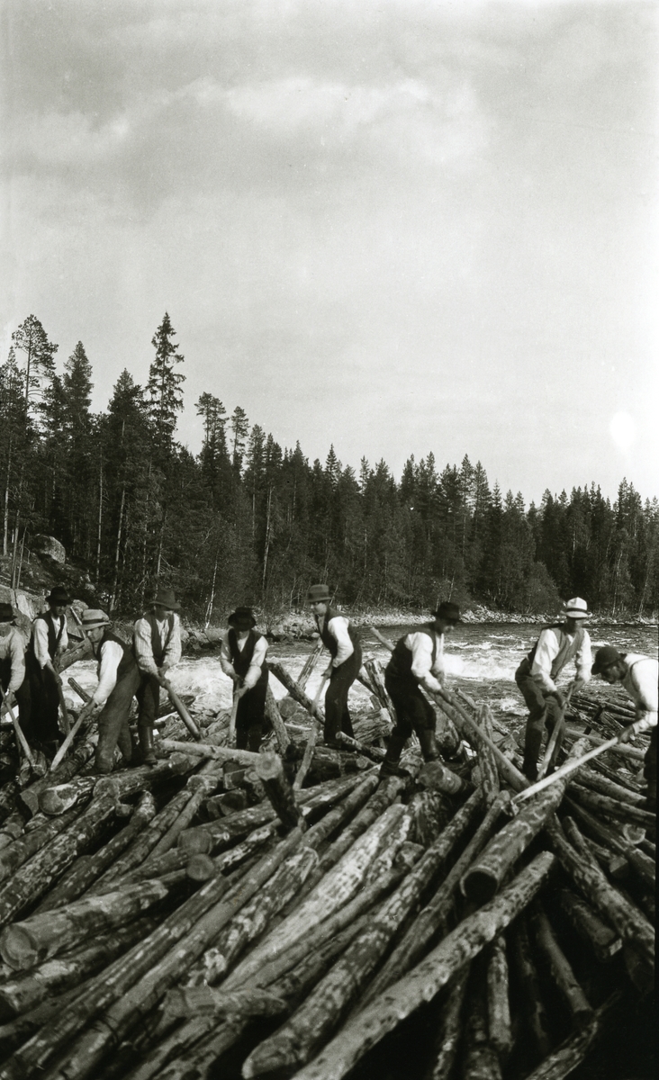Menn, fløtere, stående midt i fossestryk oppå en enorm haug med tømmer. I Sandkjellfossen, riksgrensa.