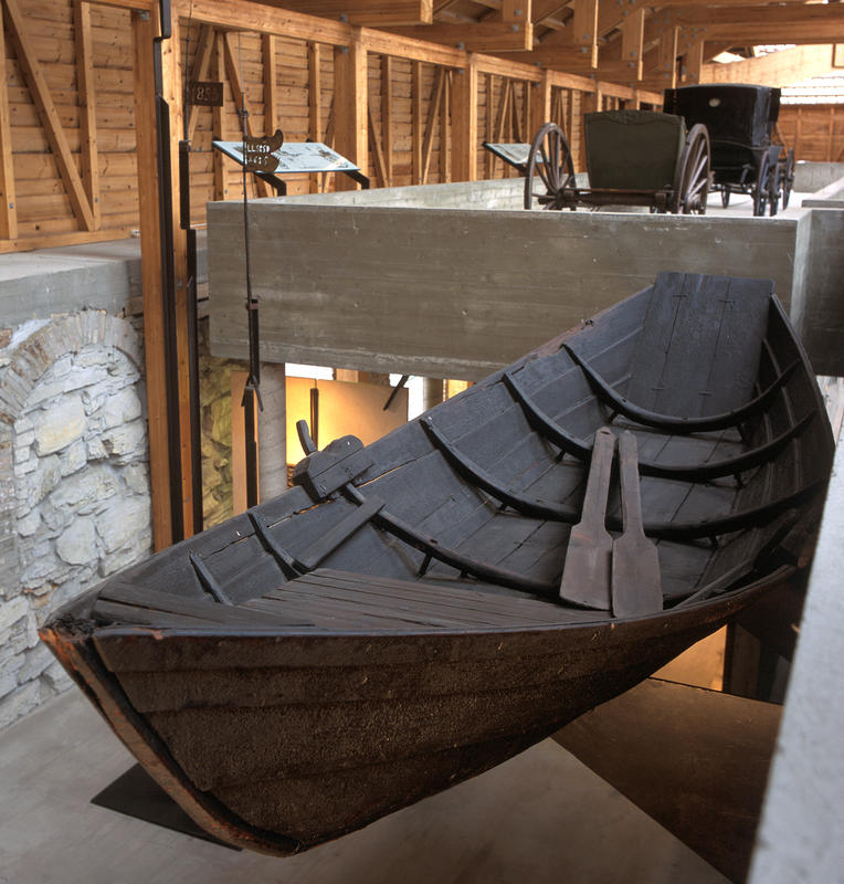 Mørkebrun robåt i tre henger oppe i utstilling i Storhamalåven, et par årer hviler i bunnen av båten. (Foto/Photo)