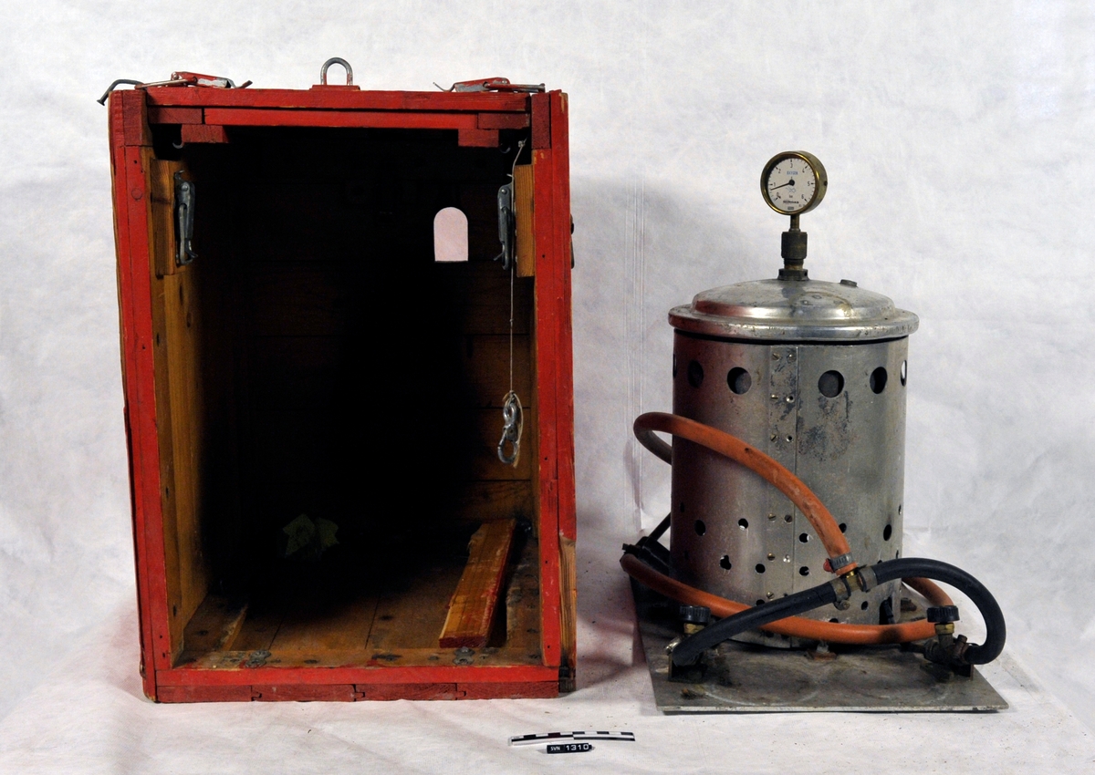 Rød kasse med sort skrift.
Kassen (A) inneholder et aparat (B) med trykkmåler og slanger.
Aparatets funksjon er usikker.