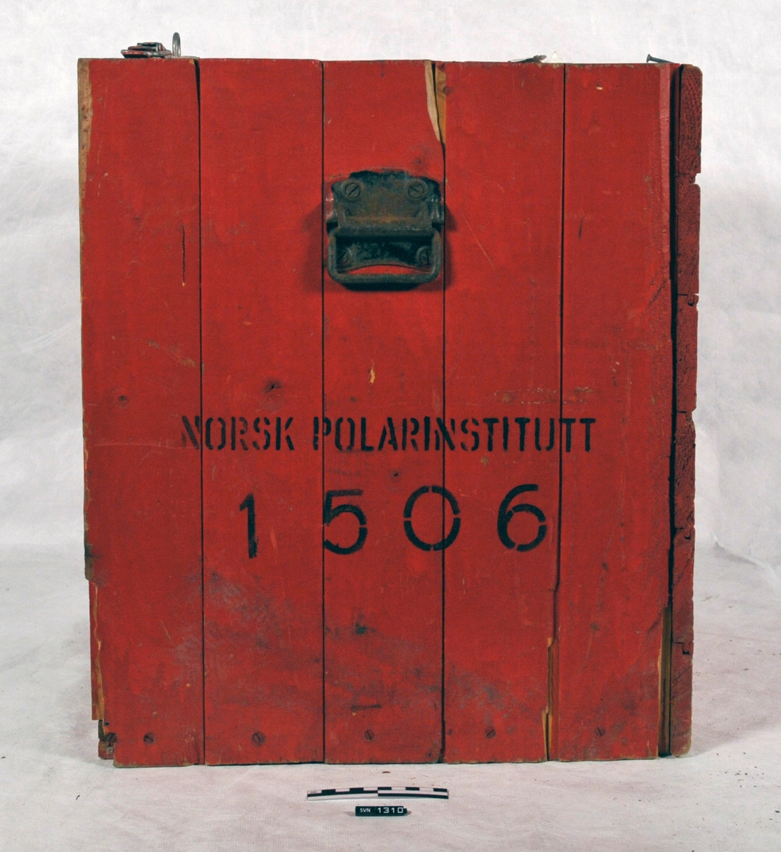 Rød kasse med sort skrift.
Kassen (A) inneholder et aparat (B) med trykkmåler og slanger.
Aparatets funksjon er usikker.