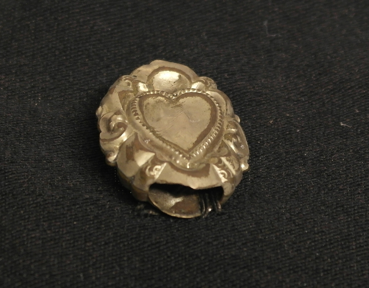 Smykkedel i gullblikk. Gjenstanden er hul og satt sammen av to like deler. Det er påført et ornament på begge sider som viser et hjerte med en ranke rundt. Gjenstanden har antakelig vært en del av et større smykke.