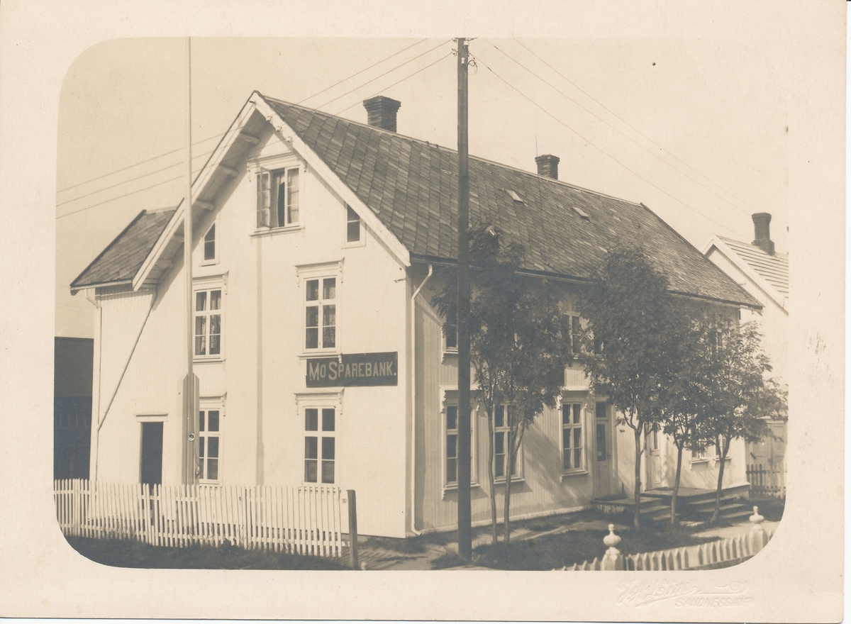 Hvit trebygning med stakittgjerde og trær foran. Skilt med Mo Sparebank.  Mo Sparebank opprettet 1. april 1876 i Mo i Rana.