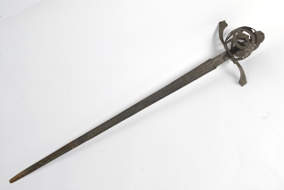 Rund pæreformet knapp med 10 fasetter  Tregrep. Flate svakt opp og nedbøyde  parerstenger med som ender i to fliker. Punslet dekor på parerstengene.
Asymetrisk kurvfeste med nyreformet parerplate på innsiden Prereplaten på utsiden er borte. Sidebøyler og håmdbøyle er formet av flate bånd. Skade på bånd på utsiden.
Tveegget rett klinge med  ovaklt  tverrsnitt  og to korte blodrender. Klingen har innskripsjon "SAHGON"