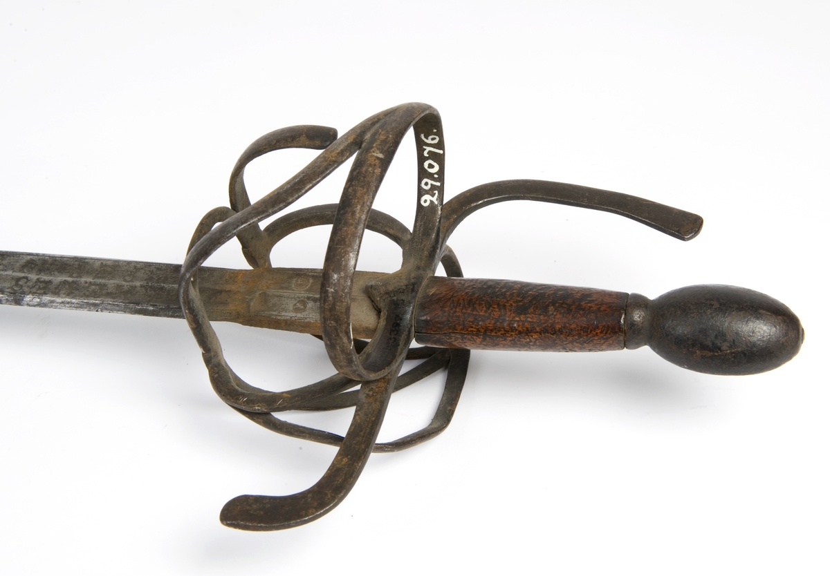 Avlang plommeformet knapp. Tregrep. Asymmetrisk feste av flate bøyler. Fremre parerstang går over i håndbøylen. Bakre parerstang nedbøyd. Sidering på utsiden med tersbøyler. Tre sidebøyler på innsiden.
Tveegget rett klinge med 6-kantett tverrsnitt og 2 korte blodrender merket MEVES / PERNS på begge sider. Klingen har 2 smedstempler på fortens fremside. Klingen er noe avkortet.