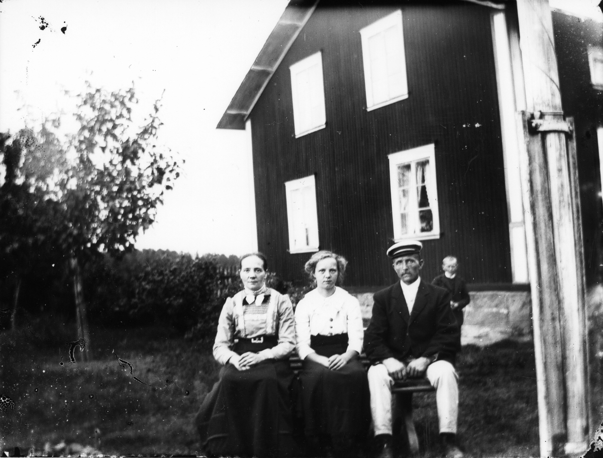 Tre äldre och en liten gosse framför husgavel.
