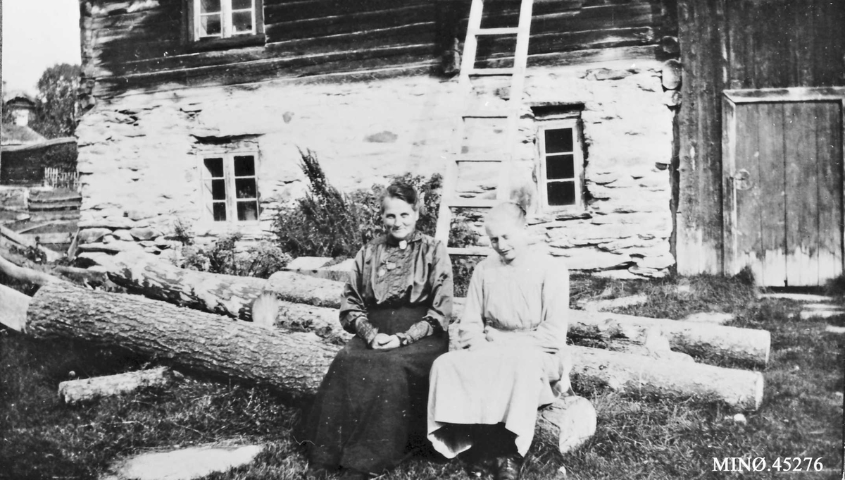 To kvinner sitter på en tømmerstokk foran fjøsbygning