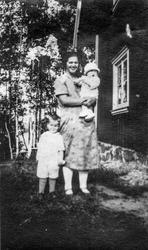 Gruppeportrett av ukjent kvinne og barn utenfor hus i skogen