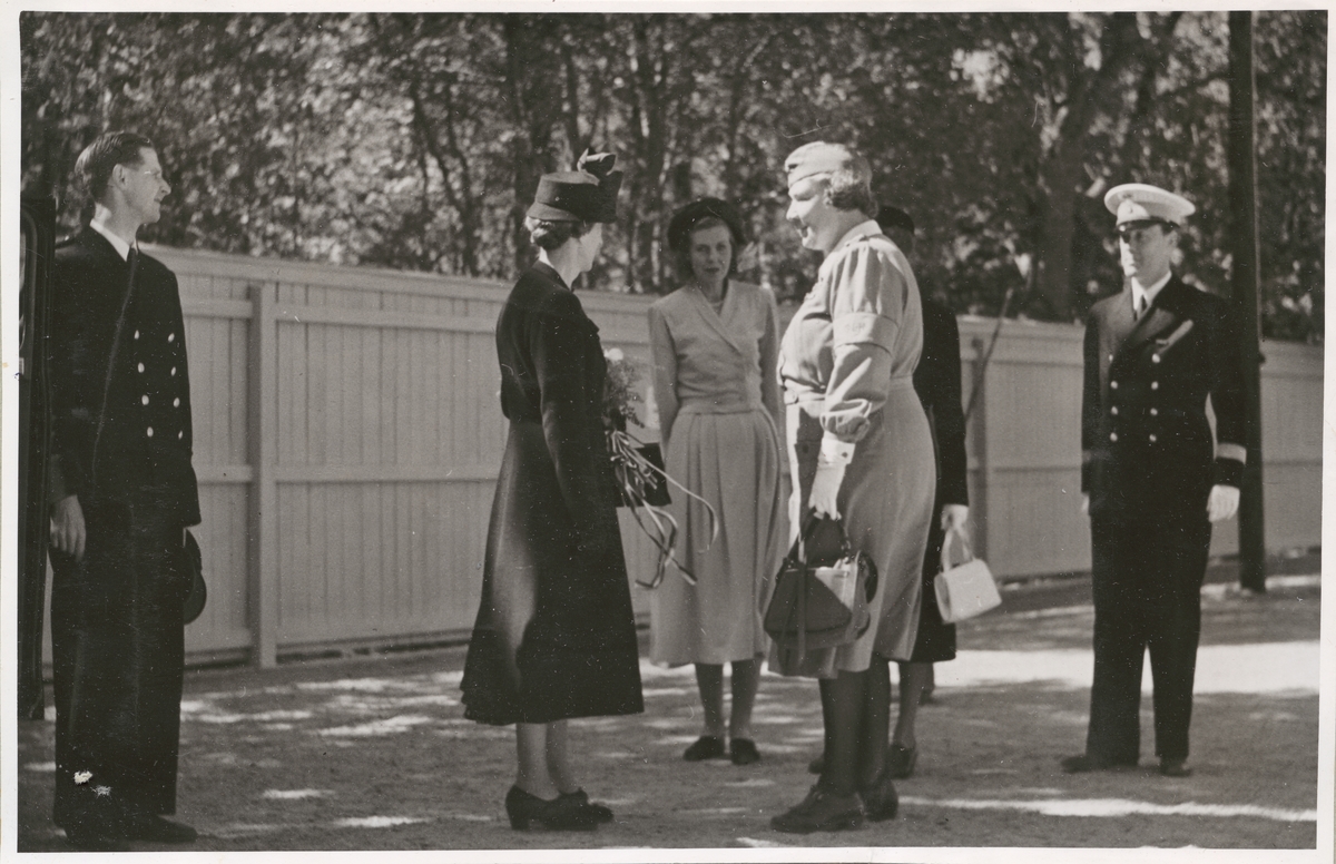 Drottning Louise med lottor och militärer på en grusplan intill ett plank. Ytterligare en kvinna finns närvarande, civilt uppklädd.