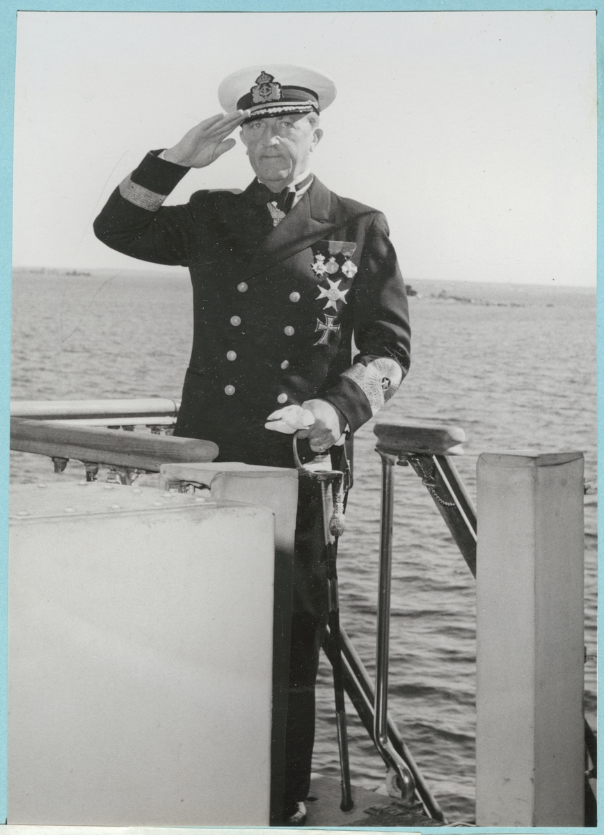 Konteramiral Samuelson går ombord på H.M.S. Montclare. Han står på däck och gör honnörhälsning med skärgården i bakgrunden. Från Engelska besöket i Karlskrona den 9-14 september 1951.
