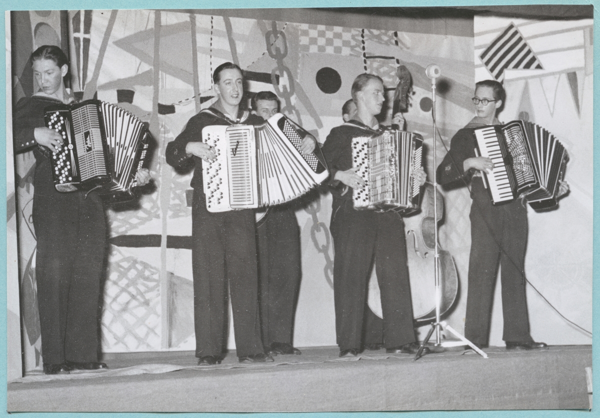 Sex uniformsklädda flottister står på en scen och spelar på musikinstrument, bl. a. dragspel och ståbas. Framför dem står en mikrofon på ett stativ.