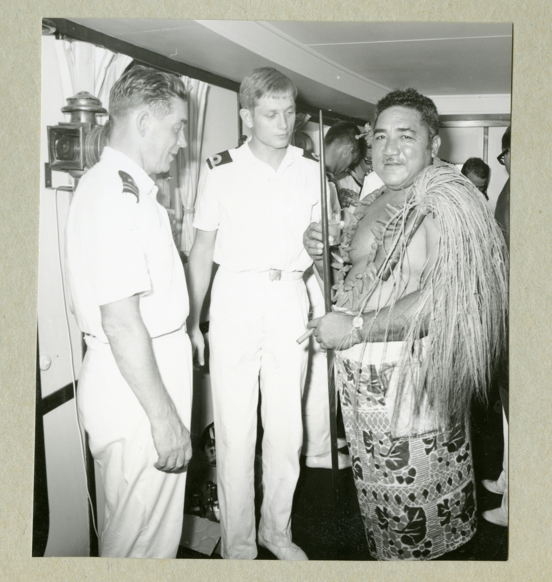 Bilden föreställer två besättningsmän i vita uniformer som står tillsammans med en man i lokala kläder. Bilden är från en mottagning ombord i Älvsnabbens gunrum för lokala dignitärer i Pago-Pago, Samoa. I mitten syns fartygets 2. fartygsintendent (FP2), fänrik Peder Ribbing.