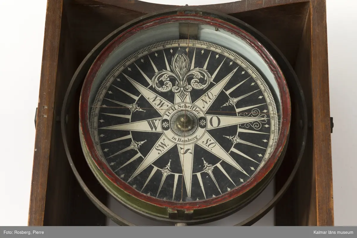 KLM 13605. Kompass, skeppskompass. Kompassen hänger i låda av ek med upplyftbart lock. Signerad på kompassrosen, J.C.H. Scheffler in Hamburg. På lådlockets insida skrivet, Skonert Augusta 1852 samt initialerna ACST.