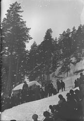 Tilskuere langs hoppkanten under skihopp. Hopper i lufta.