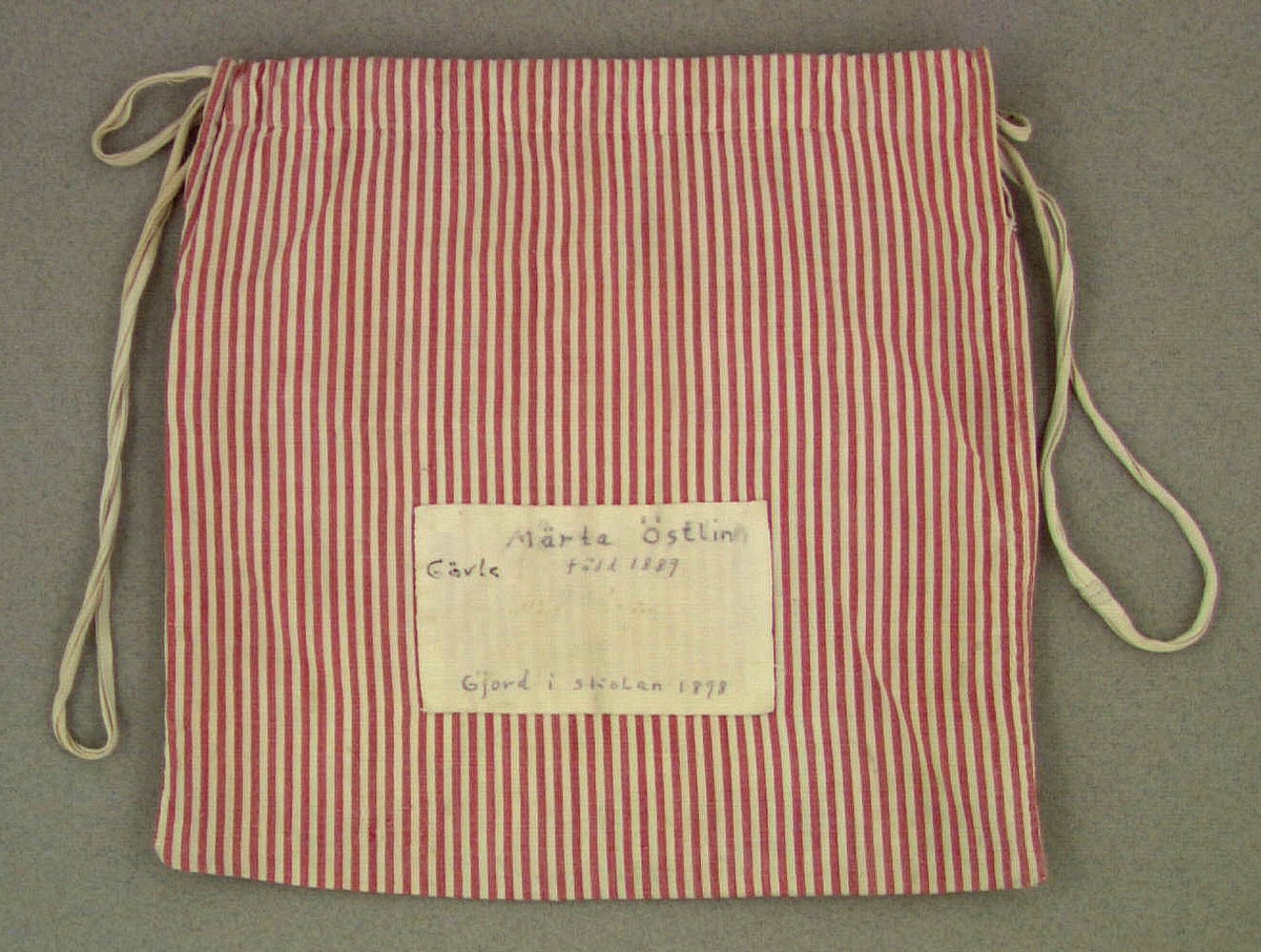 Handsydd tygpåse av röd, vitrandigt bomullstyg, maskinvävt tuskaftat. Upptill en dragsko med ett vit bomullsband. På ena sidan finns en vit tyglapp med texten: Märta Östlin Gävle, född 1889 gjord i skolan 1898.