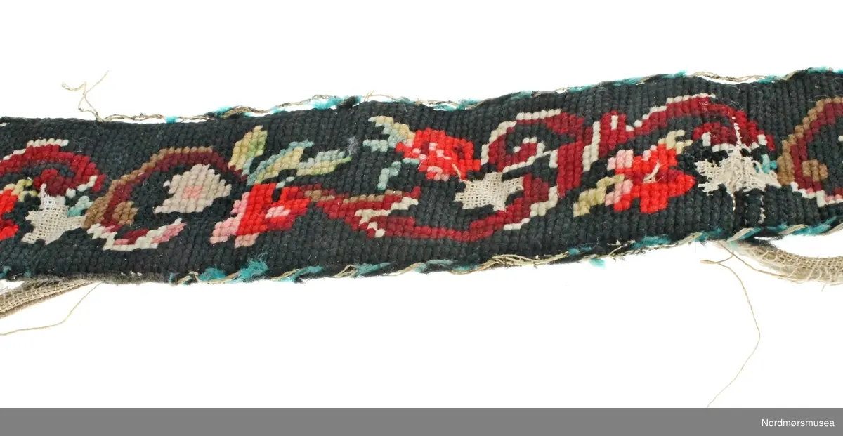 Mannsbelte brodert på stramei med blomster- og bladmotiv i mange farger. Beltet er fôret med svart ullstoff og er påsydd ei sølvfarga metallspenne. Innleggsfor i lin. Beltet er svært slitt og fôret er revnet.