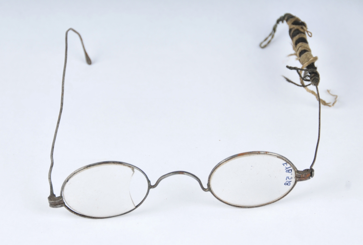 Nikkelbelagte briller med glas. Halvmaskinelt utført. Ei av stengene er reparert med messingstråd som det er surra ein bomullstråd rundt.