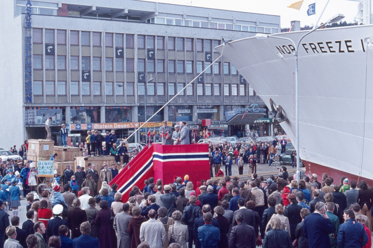 Folkemengde på kaia i anledning skipsdåp. Flaggdekket podium midt i bildet, Rasch-Tellefsenbygget i bakgrunnen.