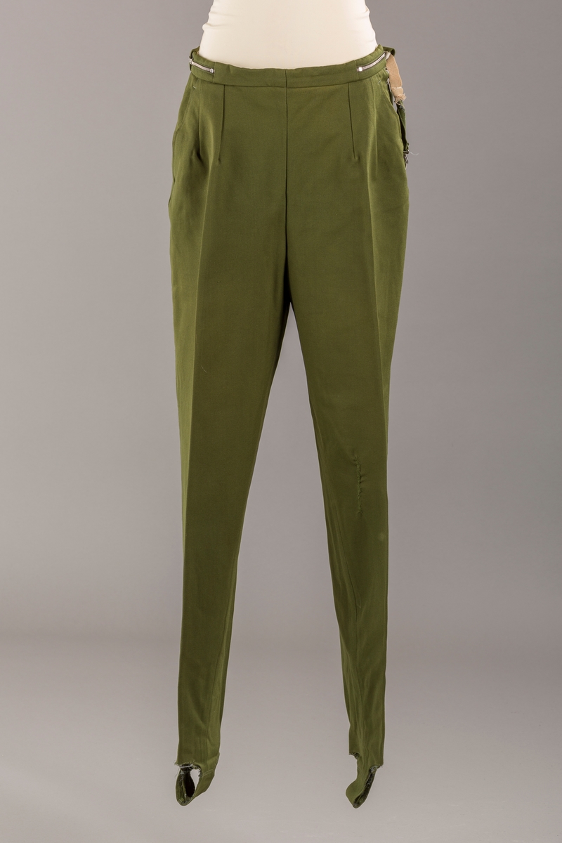 Grønn strekkbukse med stropper under bena. Avsmalnende ben. Glidelås i venstre side. Elastisk spennglidelås.