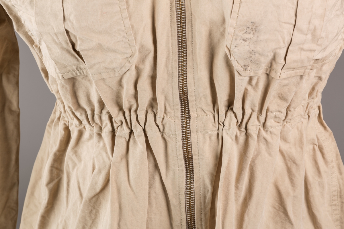 Form: Vanlig anorakkfasong med strikk i livet. 2 brystlommer med knapp.
Hette med glidelås på topp midt, snøring.
Glidelås helt ned foran.
Innermansjett med strikk i ermene.