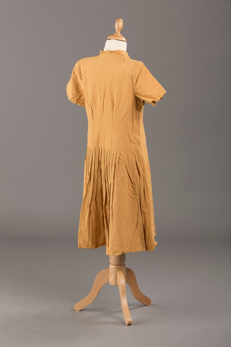 Form: Korte ermer, livet rynket til bærestykke foran, plissert skjørt, tre pynteknapper, en trykknapp, forlomme.