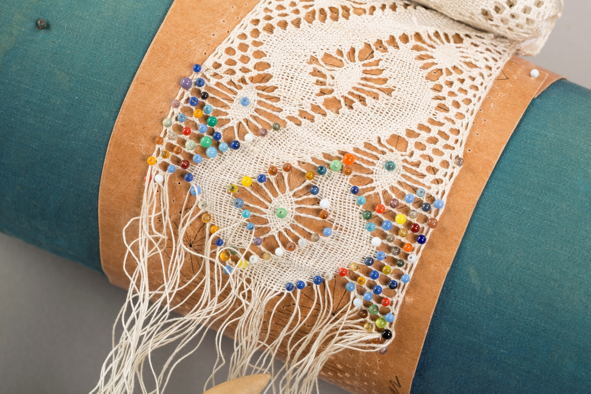 Form: Sylinderformet "pute" hvor stivt pappstykke rundt puta viser mønsteret. 
Kniplingsstykke under arbeid påspent med knappenåler
Pinner medkniplingstråd